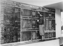 Titel: Vertrekbord in stationsgebouw met erop vermeld vele legendarische vliegers van de KLM Vluchten werden toen met de hand met krijt op het planbord geschreven (20 08 1931) Op het linkerbord staat 2e van onderen de 42e Indie-vlucht met de PH_AFS aangegeven., — Photo Amsterdam City Archives / Unsplash