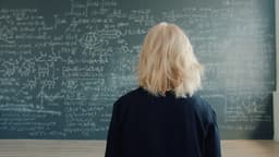 Female scientist is walking to chalkboard wall with formulas in university classroom thinking about scientific project. People and lifestyle concept. — Photo Vitaly Gariev / Unsplash
