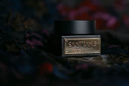 A closeup of an eye cream product called Saint Jane, Bright Repair Eye Cream, that is made from Vitamin C, Green Tea and CBD on a dark black and floral background with luxury, high end black and gold packaging. — Photo Kitera Dent / Unsplash
