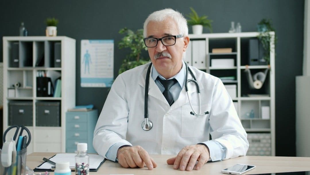 Portrait of joyful doctor senior man in uniform talking waving hand showing bottles with pills in hospital office during online call with patient. Medicine, people and consultation concept. — Photo Vitaly Gariev / Unsplash
