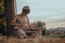 woman wearing beige blouse and pants sititng on brown grass field bihind of tree bark while playing musical instrument — Photo Michael Walk / Unsplash