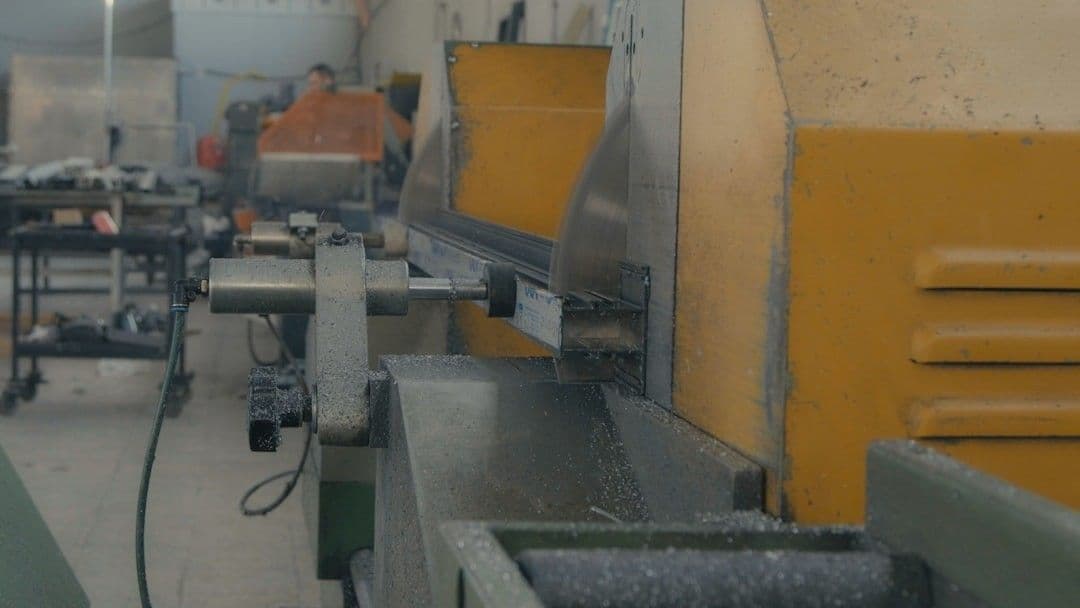 A high-speed industrial saw cuts through a metal piece, sending fine metal shavings into the air. The motion blur on the spinning blade emphasizes the power and precision of the cutting process. — Photo Cemrecan Yurtman / Unsplash